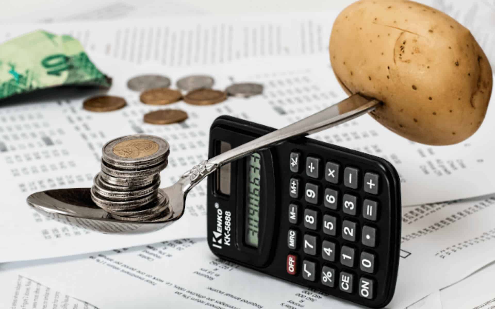 A spoon with coins in the bowl and a potato on the other end is balanced on a calculator while it all sets upon printed financial papers
